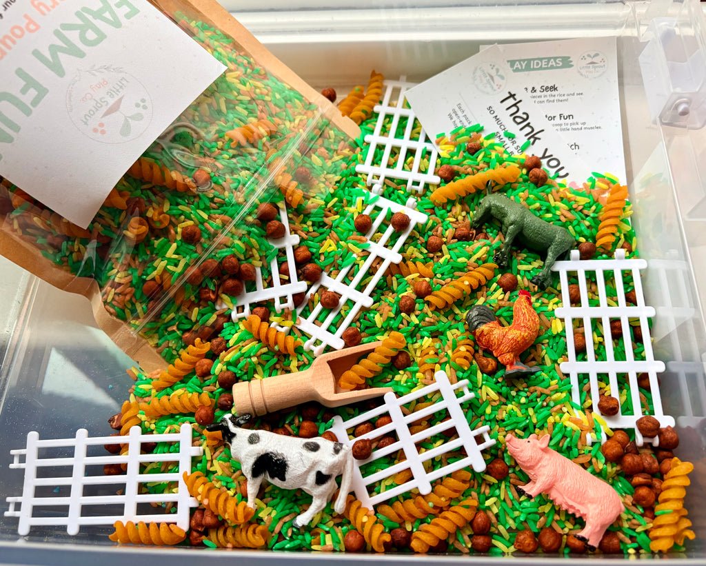 A clear plastic sensory bin filled with green and orange spiral pasta, white fence pieces, small brown beads, and farm-themed toy animals including a cow, pig, chicken, and horse. A brown paper bag labeled Farm Fun is tipped into the bin, along with printed cards, in a bright studio-style setup.