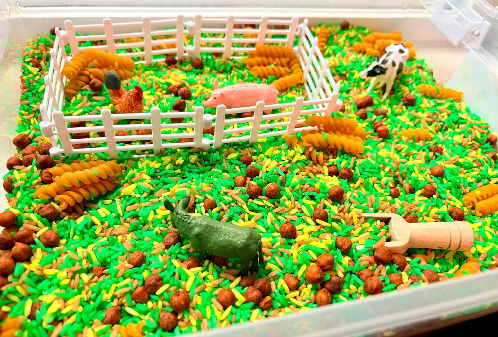 A children's sensory play bin filled with bright green and yellow rice, brown chickpeas, a white plastic fence, toy animals including a cow, pig, chicken, and donkey, and a wooden scoop.