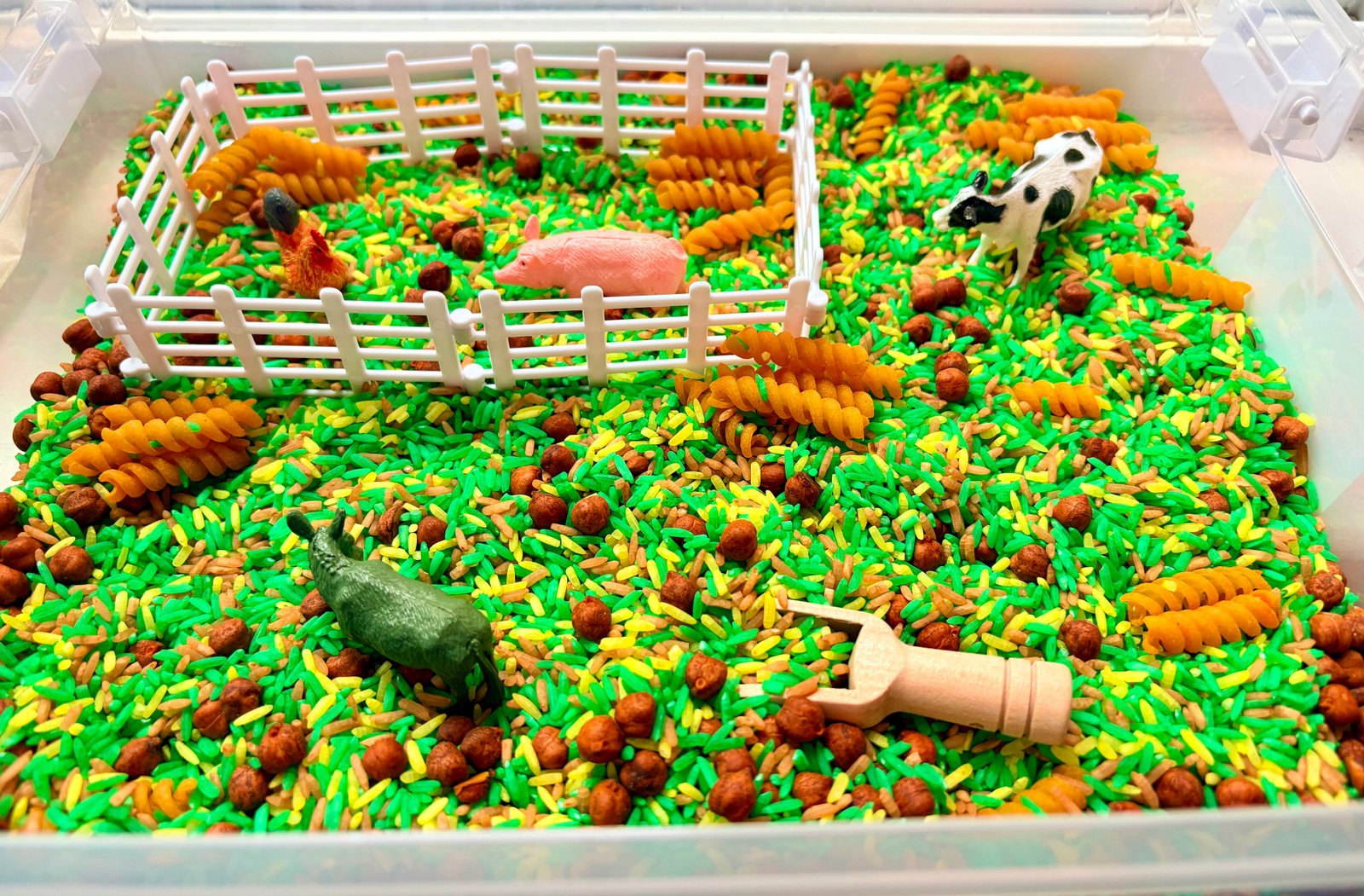 A children's sensory bin filled with bright green and yellow rice, brown pellets, yellow pasta resembling hay, a white plastic fence, a pink pig figure, a black-and-white cow figure.