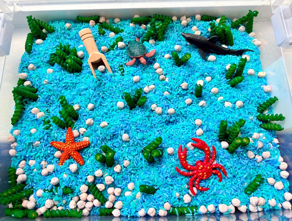 An ocean-themed sensory bin filled with bright blue rice, white pebbles, green seaweed, a wooden scoop, a shark toy, a turtle toy, an orange starfish, and a red crab toy, photographed from above inside a clear plastic tray.