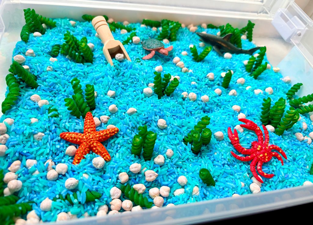 An underwater-themed sensory play box filled with bright blue rice, white pebbles, green seaweed, a wooden scoop, a gray shark toy, a green turtle, and orange and red starfish and crab figures.